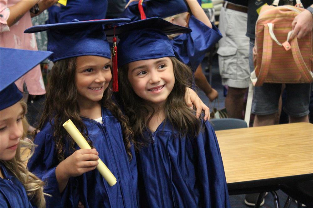 Two kindergarten girls wearing caps and gowns stand with arms around each other