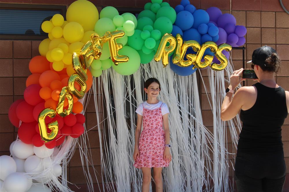 A student wearing a pink dress stands by a rainbow colored balloon arch with white streamers behind